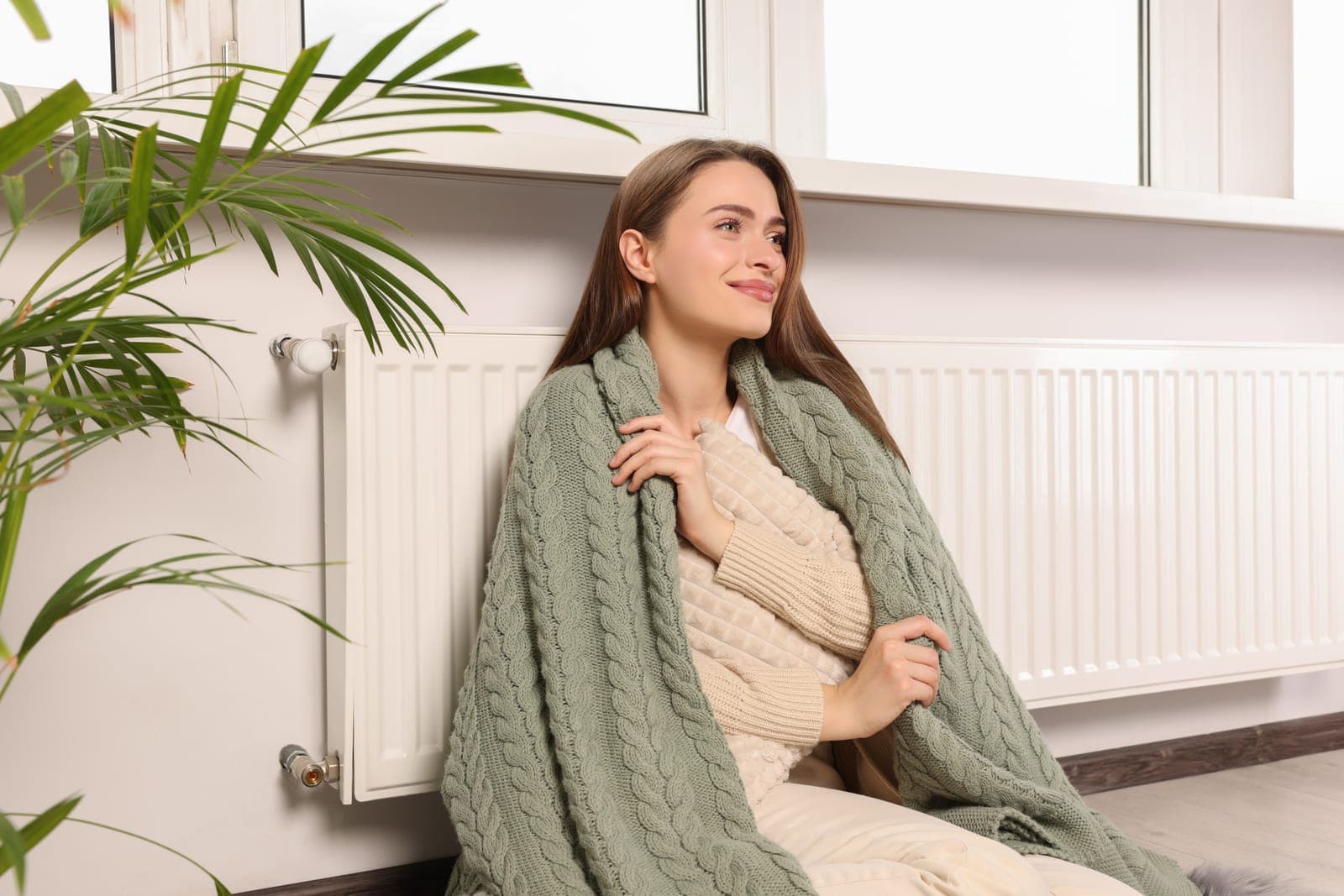 A woman wrapped in a blanket beside a radiator in a cold home