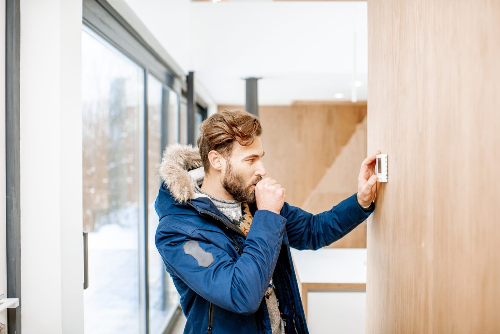 A man in a winter coat adjusting his home thermostat on a cold day